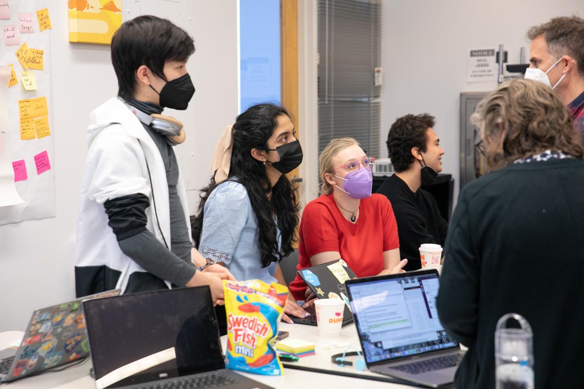 A group of three students wearing face masks talk with professors in a classroom.