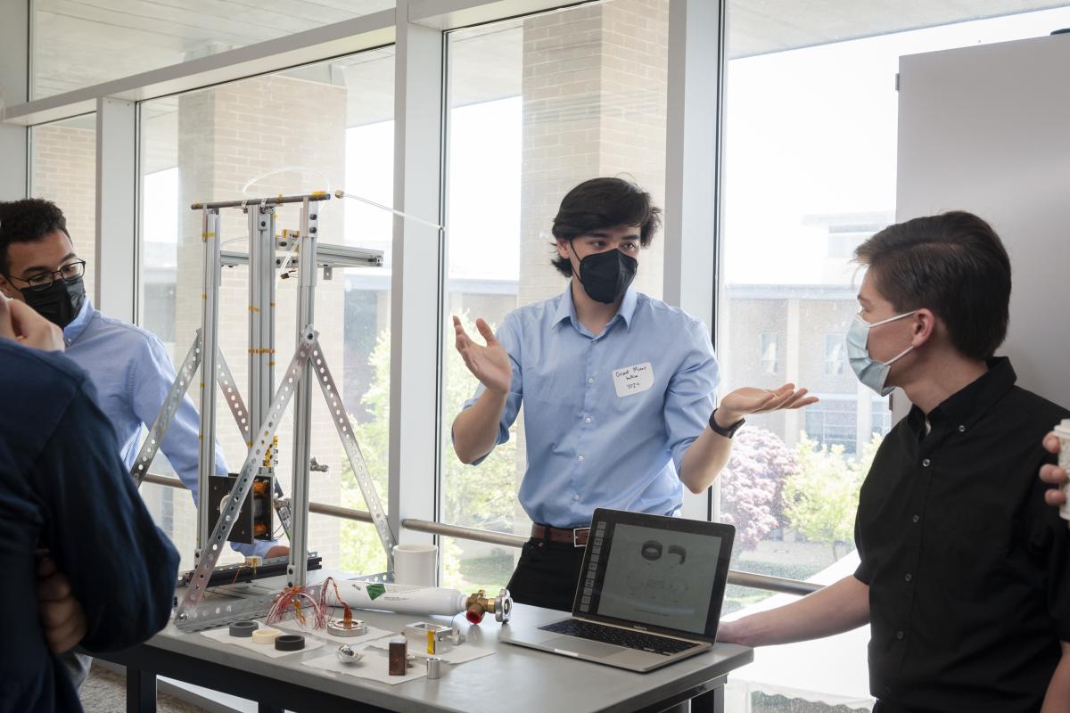 Team member Grant Miner '24 talks with Braden Oh '23 about the project at Spring Expo 2022. Three students discuss a project while behind a table that displays a laptop and metal structure (i.e. a thruster).