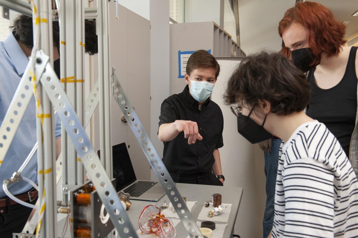Braden Oh '23 (center), points to the thruster while interacting with students and members of the Olin community during Spring Expo 2022. A student wearing a blue face mask points to something on a table, with a large silver apparatus in the foreground.