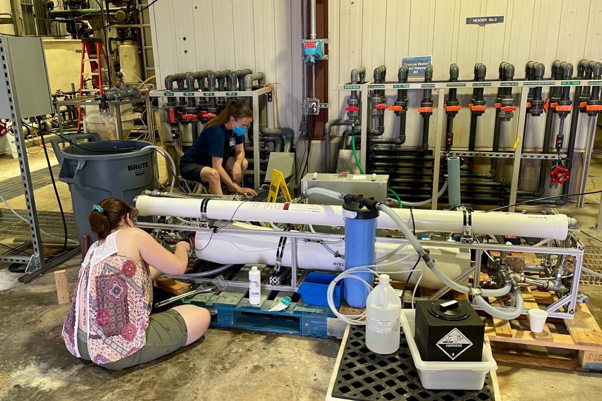 Two people are sitting cross-legged working with their hands on two large white tubes.