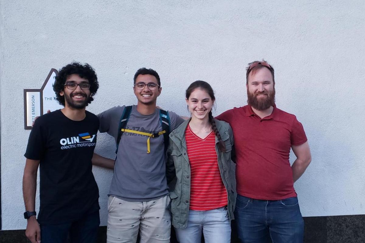 Three Olin students interning at Tesla, Inc. pose with Nick Ostrom '14 at the facility. Four people pose in front of a grey wall.