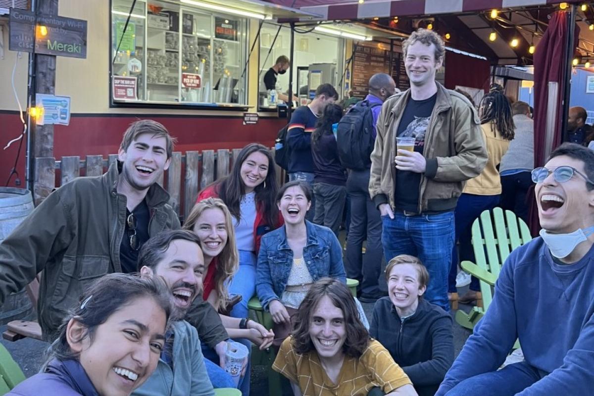 A group of ten people smile and share a laugh while gathering in front of a food truck. A group of ten people smiling and laughing in front of a food truck.
