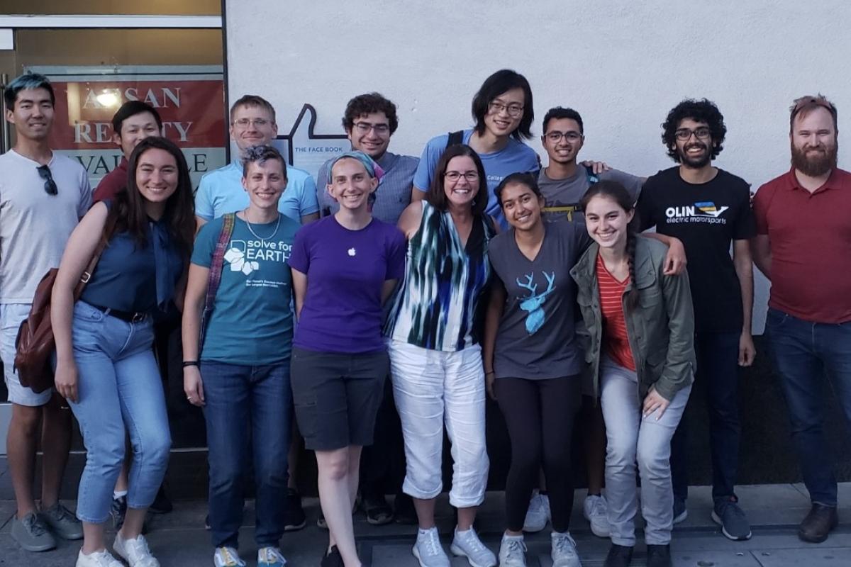 A large group of Olin College alumni and students pose with Sally Phelps, Director, for a group photo in the Bay Area. A large group of people pose in front of a white wall.