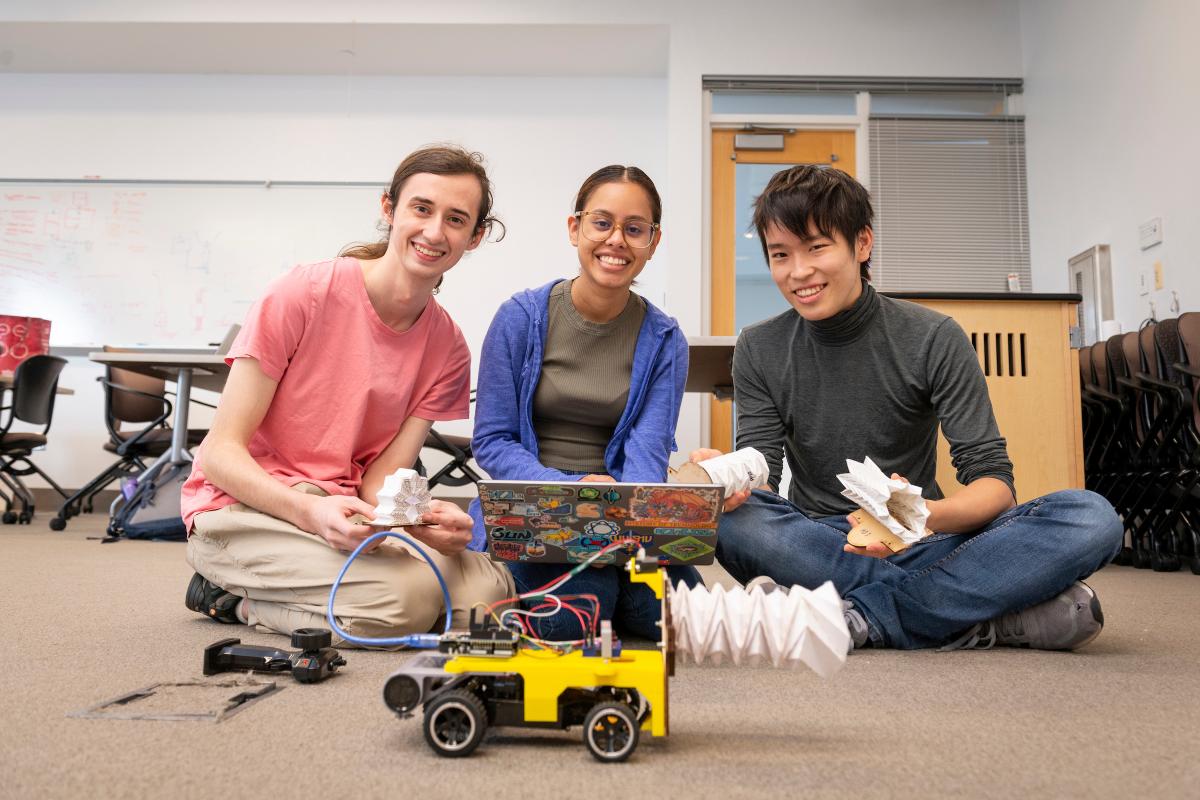 Three students sit cross-legged on the floor, looking at camera and smiling while a yellow robotic vehicle is posed in front of them.