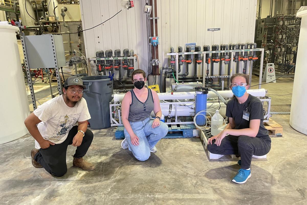 Three people squat in front of an inland water purification system prototype.