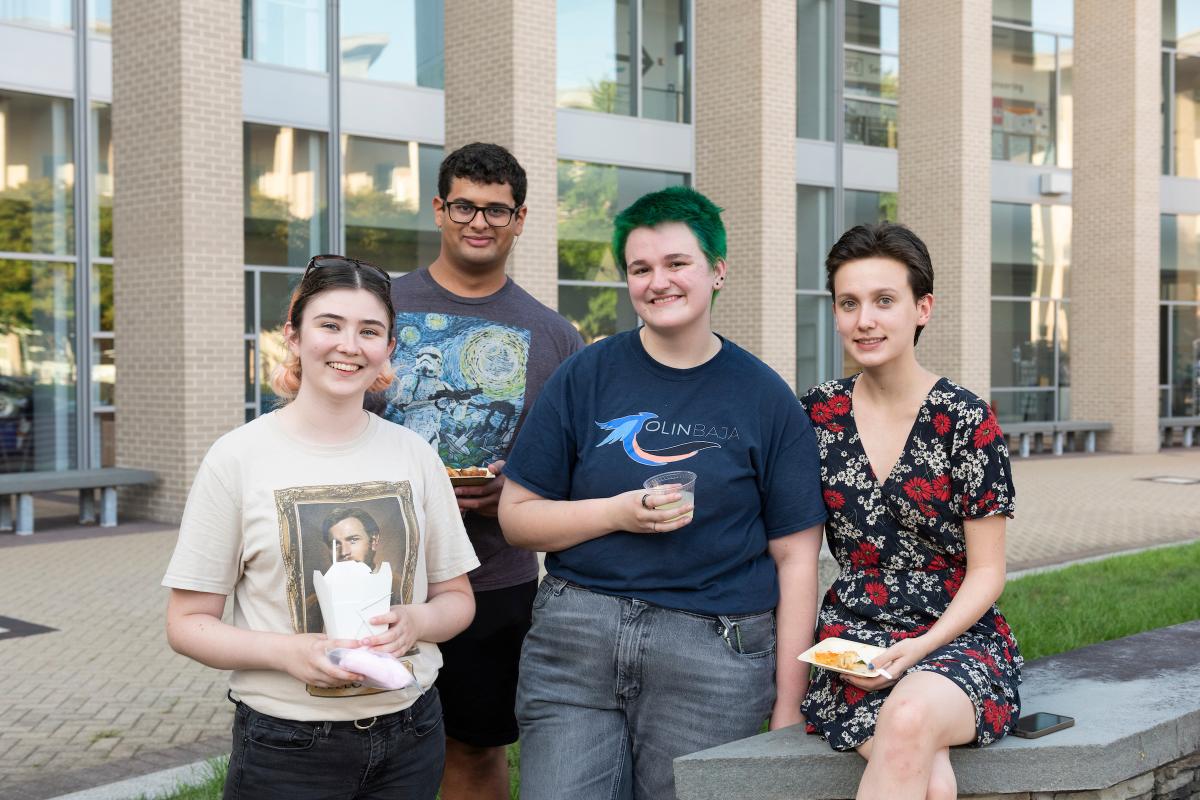 Four Olin students pose for a photo outside in front of a building with glass windows. Four students pose for a photo outside in front of a building with glass windows.