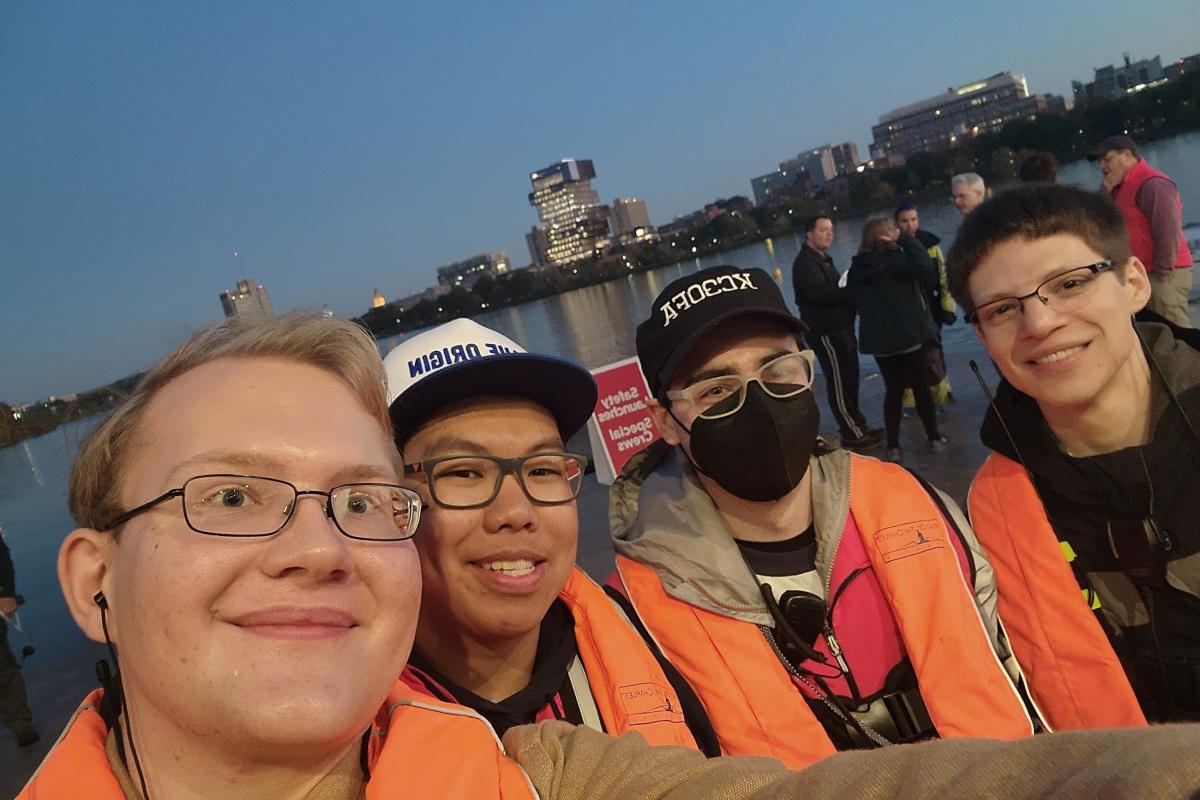 Four student-volunteers in orange life-preserver vests take a selfie outside with the Charles River in the background.