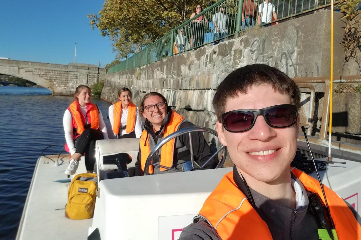 A student wearing sunglasses takes a selfie while on a motor boat. Three woman are smiling in the back of the photo.
