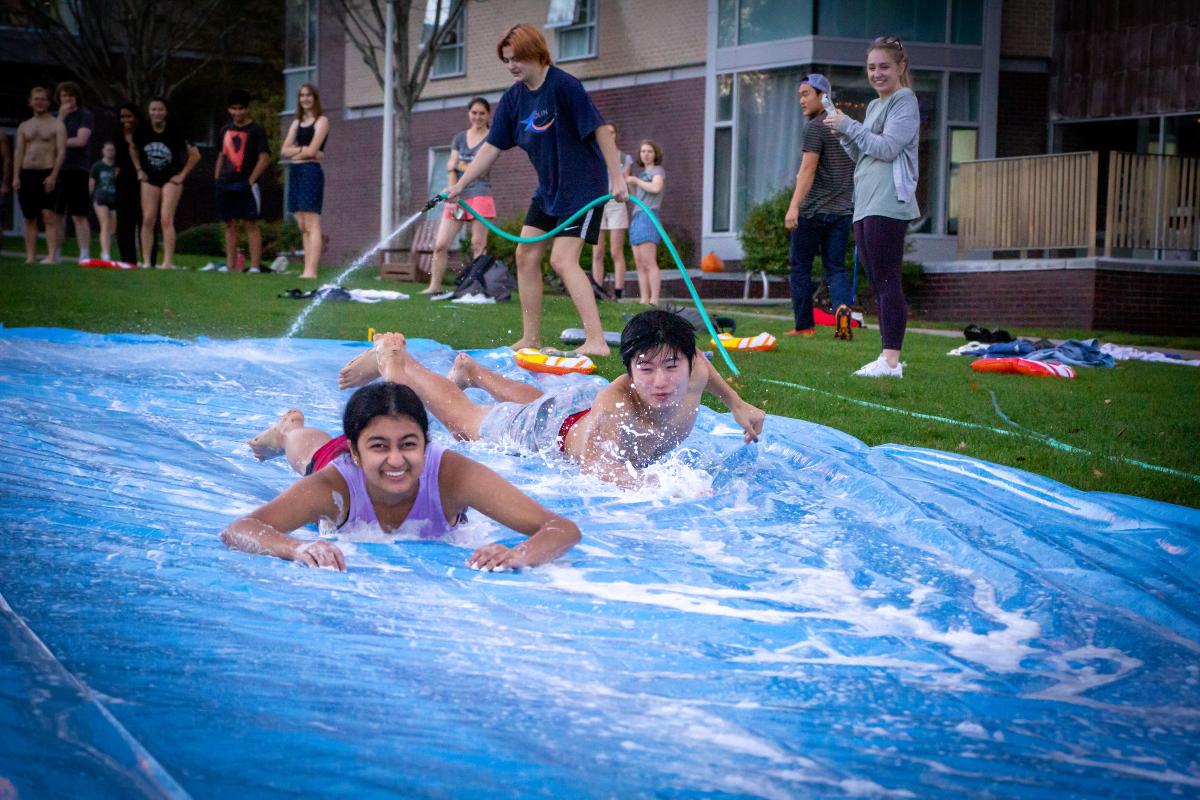 Two students slide down a water-sprayed blue slip & slide.