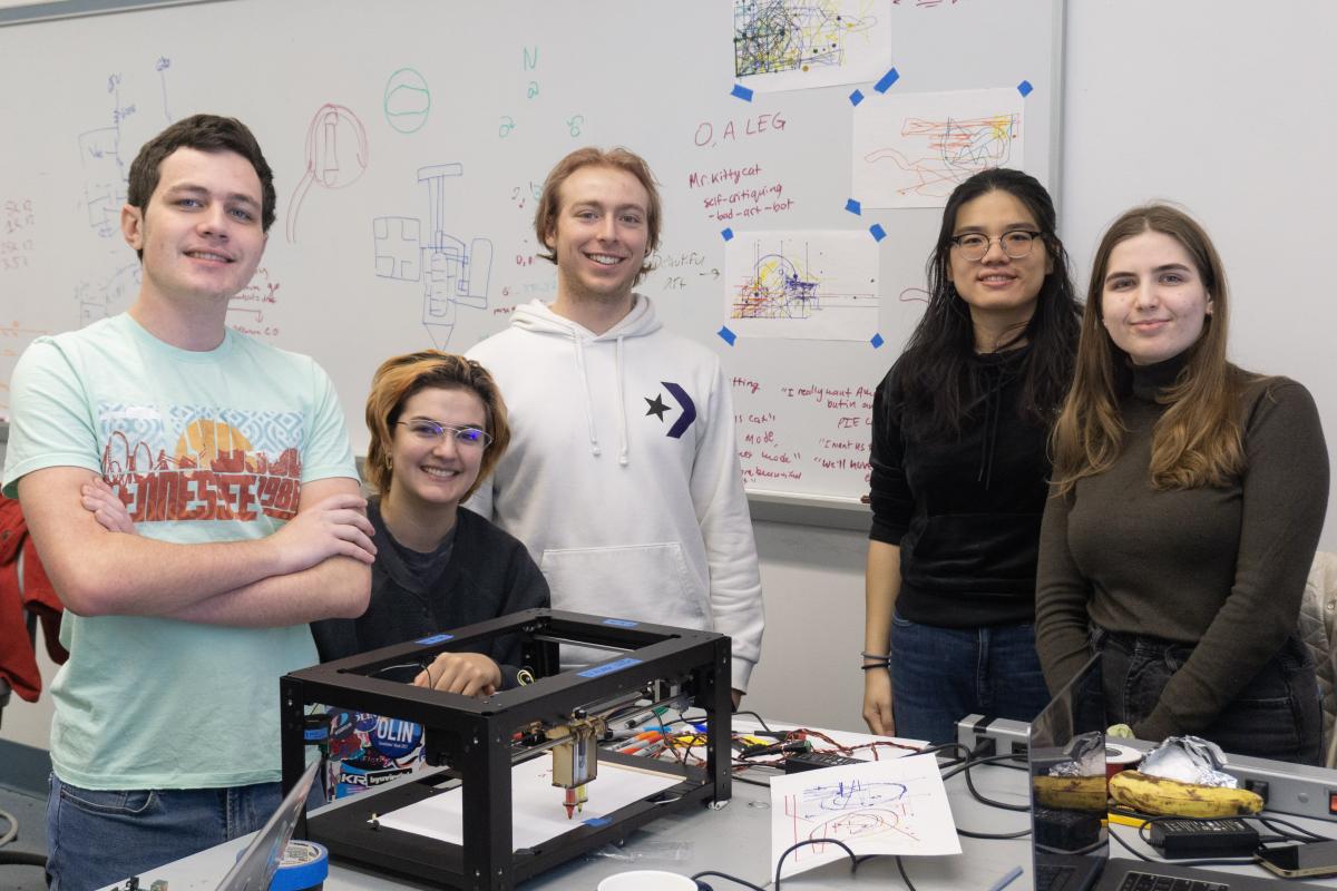 Five students in PIE Fall 2022 pose in the classroom during finals. Five students pose in a classroom in front of a rectangle box with see-through sides.