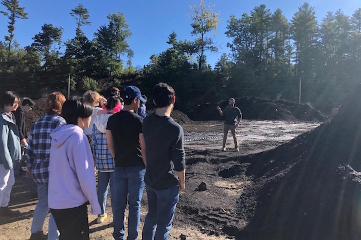 Multiple students stand outside, flanked by large piles of fertilizer-looking materials. Multiple students stand outside, flanked by large piles of fertilizer-looking materials.