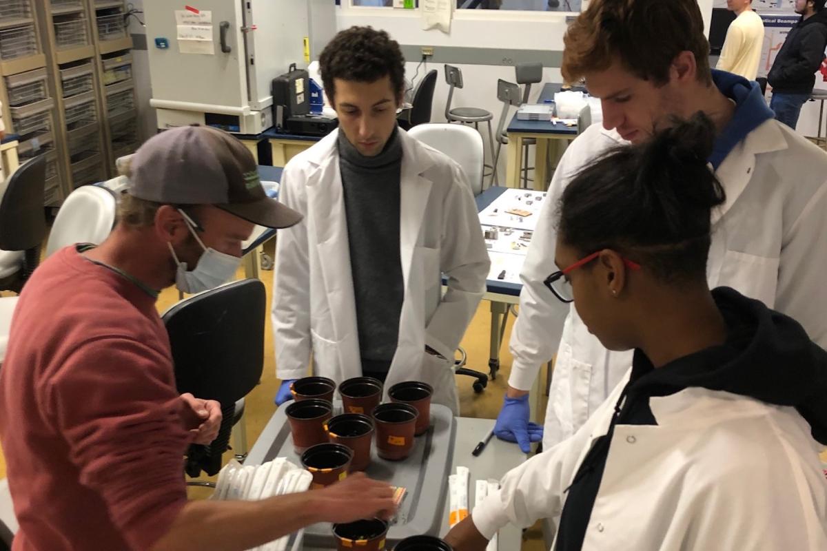 Three students in white lab coats watch as a worker from Black Earth Compost tests compost tea in little containers. Three students in white lab coats watch as a worker from Black Earth Compost tests compost tea in little containers.