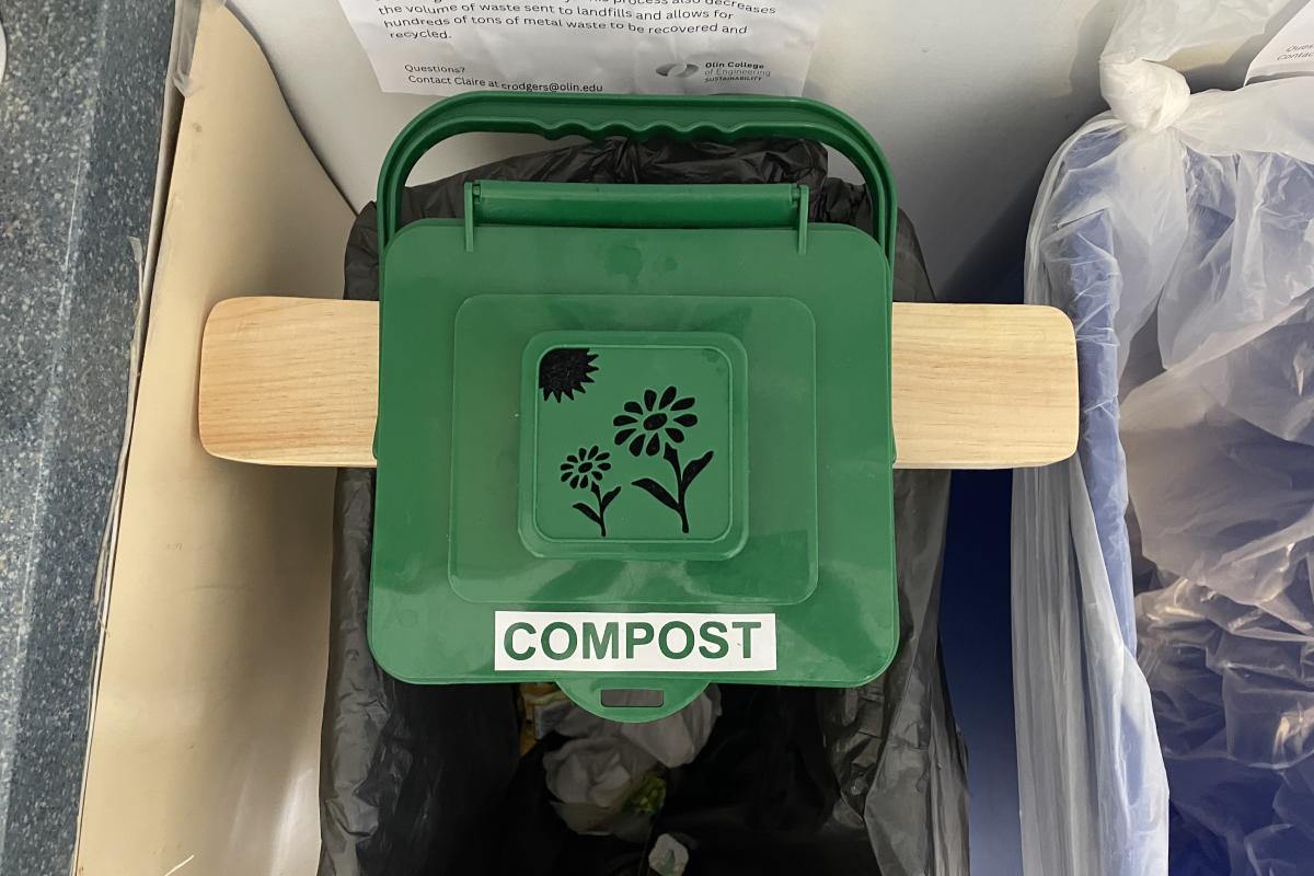 An image of the ECO class's small, green compost bin attached onto a garbage bin in Milas Hall.. An image of a small, green compost bin that attaches onto a garbage bin.