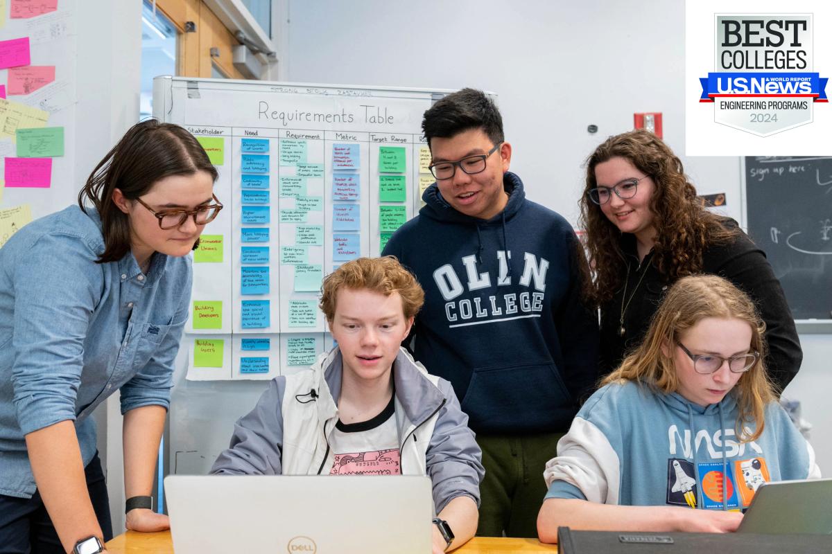 Five students work around two silver laptops in a classroom. 