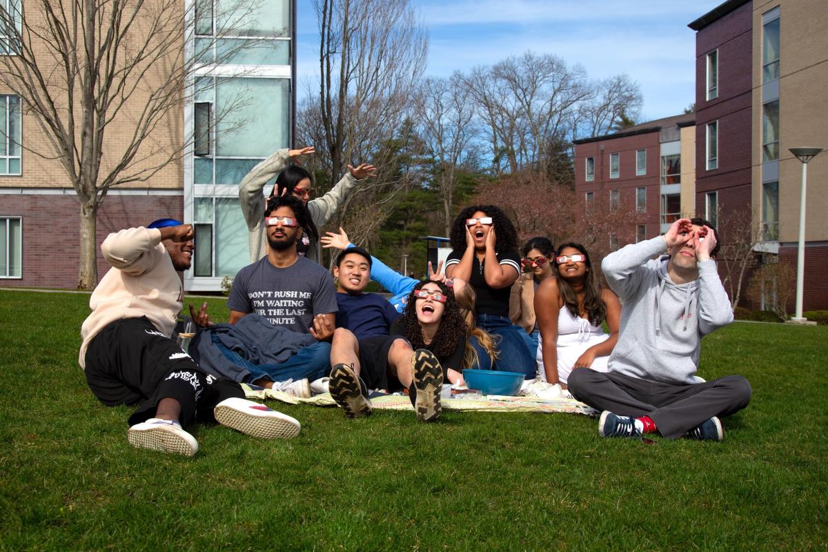 A group of young students wearing eclipse glasses pose in funny ways on a blanket while sitting outside on green grass.