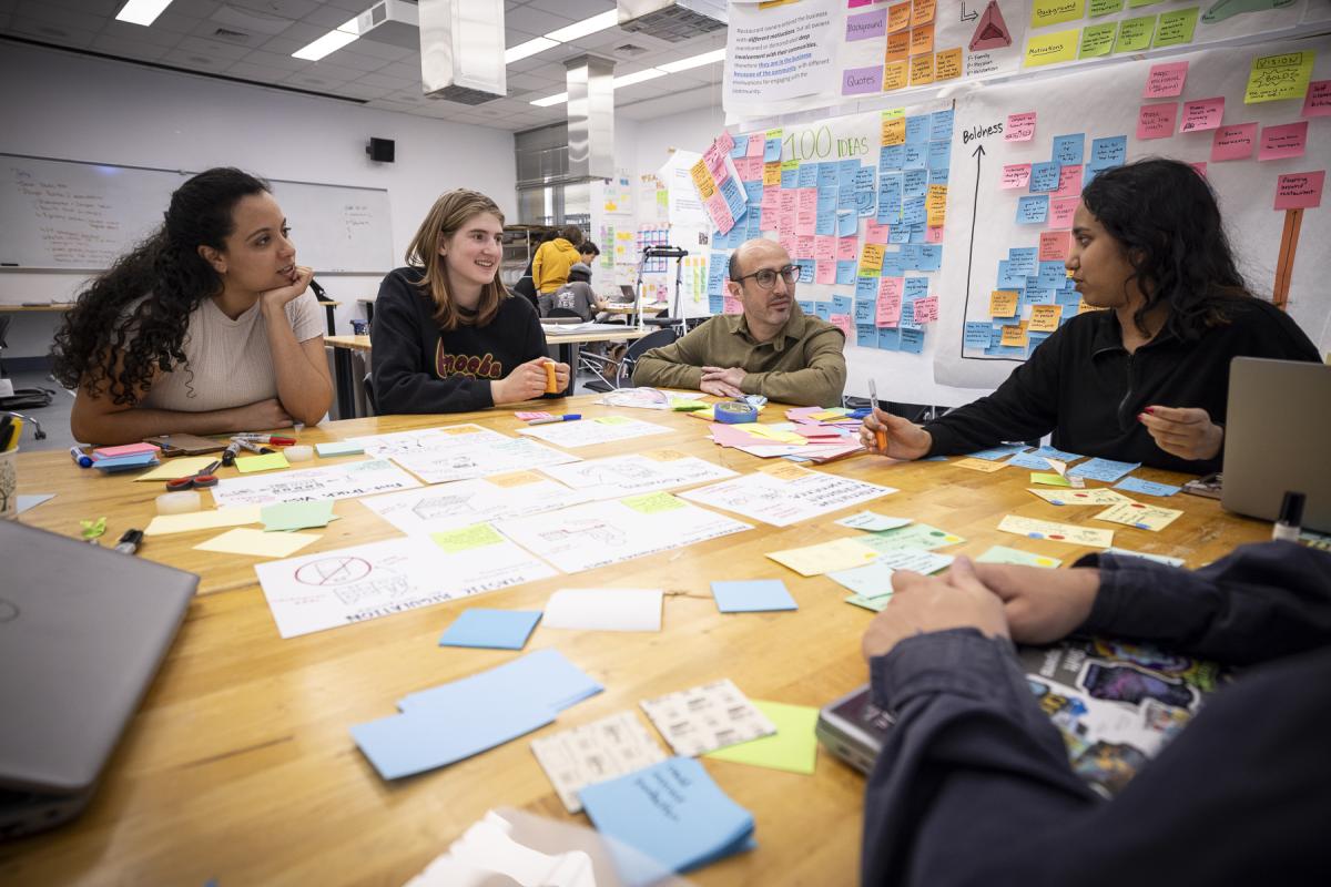 Olin students and faculty collaborate around a table in a sticky-note filled classroom. Olin students and faculty collaborate around a table in a sticky-note filled classroom.
