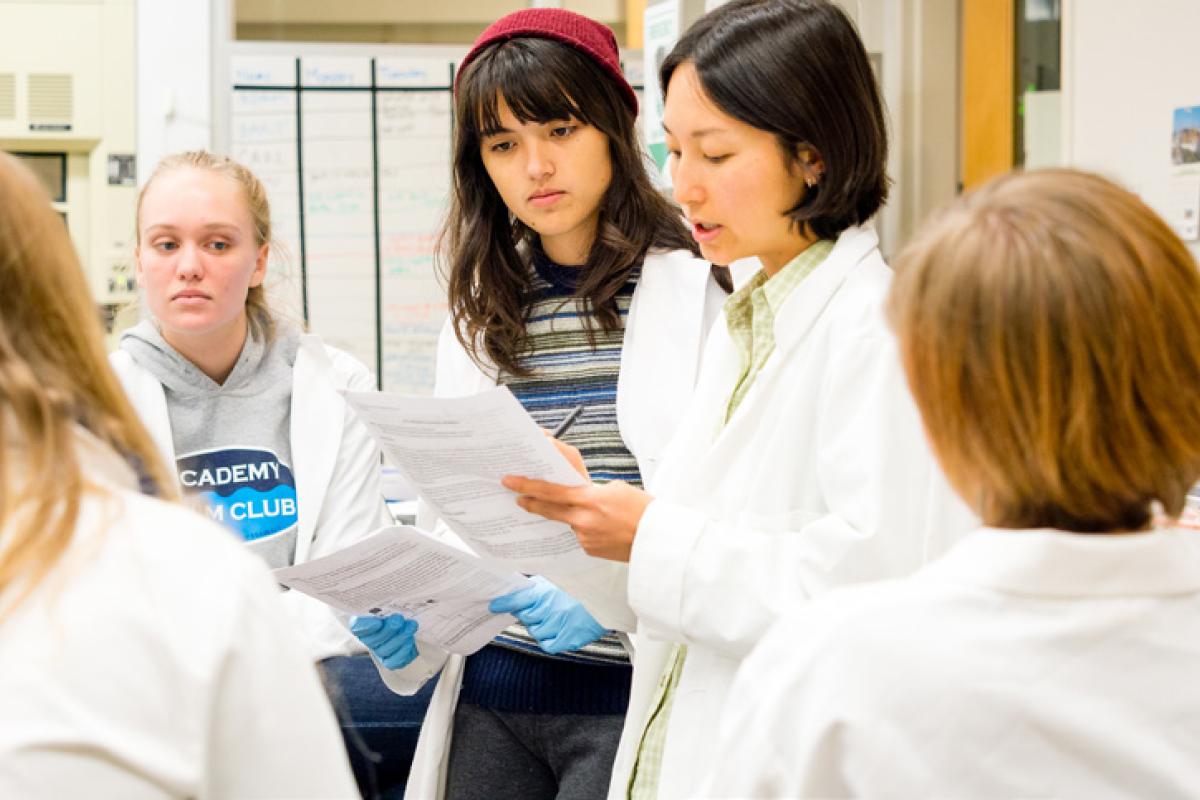 Jean Huang teaching students in a lab. Jean Huang teaching students in a lab. All of them gathered around her wearing white lab coats.