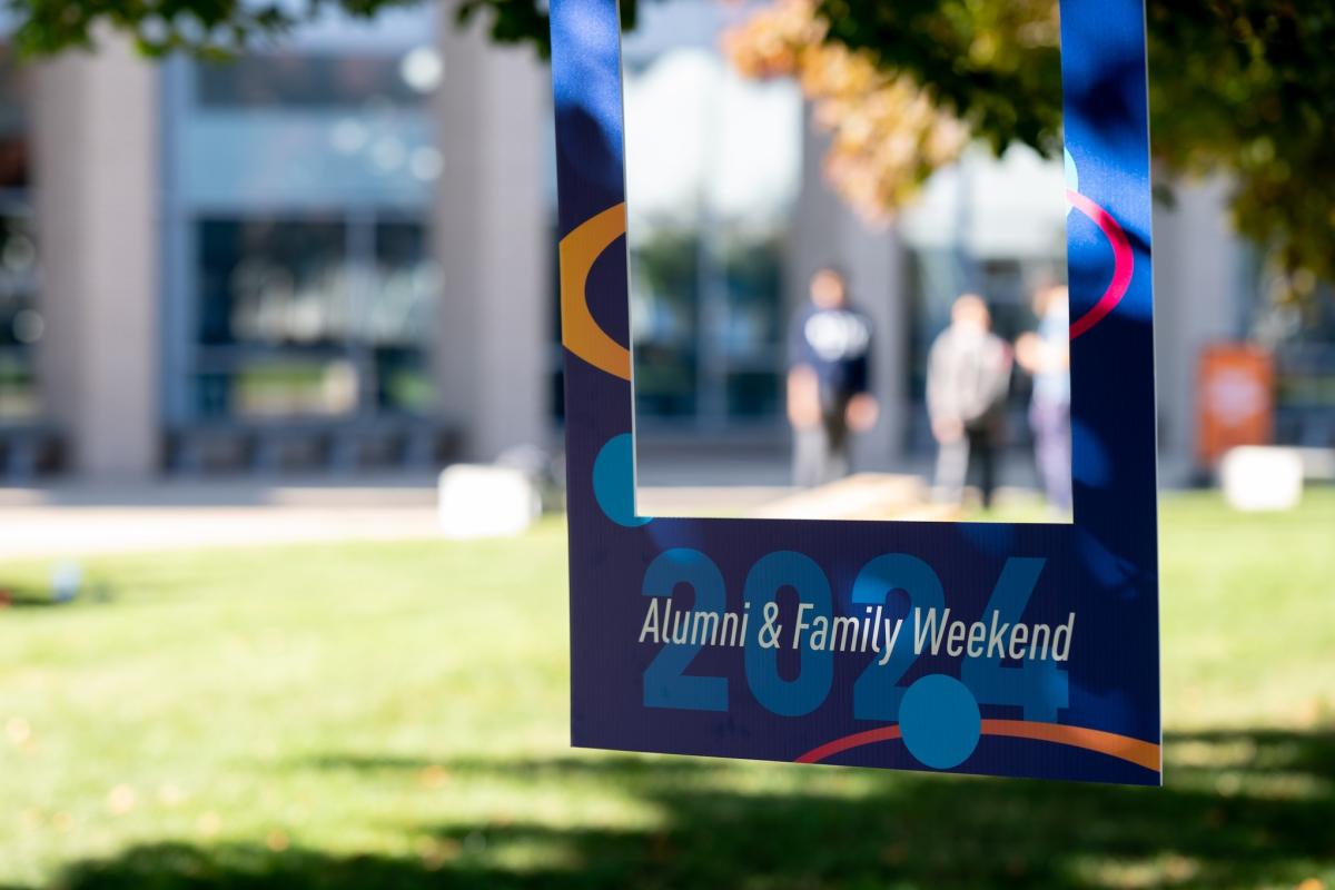 A photo frame prop with "Alumni & Family Weekend 2024" text, held outdoors with a blurred background of people and the academic building.