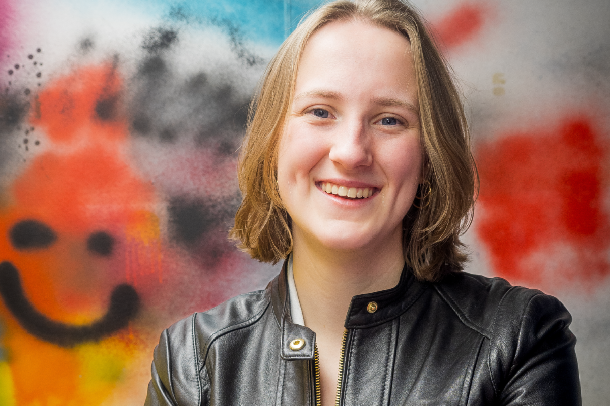 Maya McKone-Sweet'26 portrait Maya in a black leather jacket smiles confidently in front of a colorful graffiti wall.