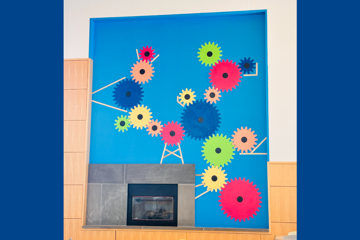 Mural Installation A colorful gear-themed mural on a bright blue wall above a fireplace, featuring interconnected wooden gears in various sizes and colors.