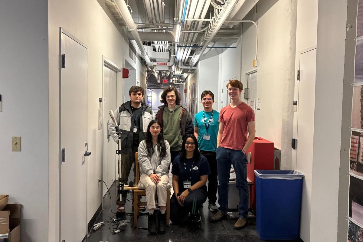 A team of six Olin students are pictured in a white hallway. Four standing and two in front sitting in chairs.