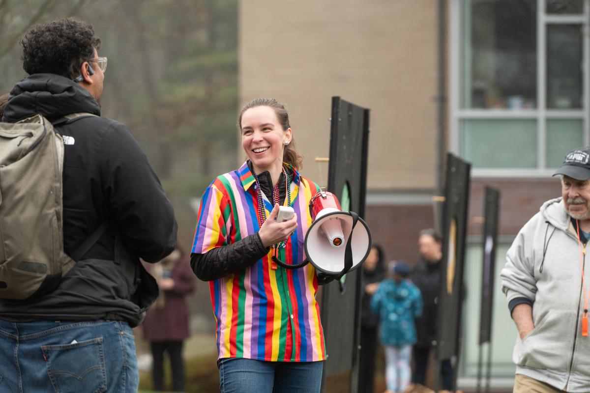 Georgia Van de Zande stand in a rainbow shirt while holding a white and red megaphone. 