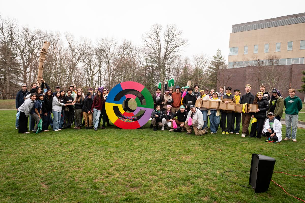 A group of six student teams stand on each side of a big colorful cylinder. 