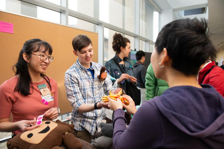 Two Olin students talk with another student during Fall Expo. Two smiling students hand another student an orange object to inspect.