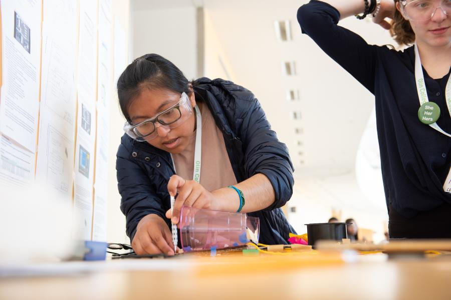 A student with safety glasses leans over a wooden table to work with a cup and a pen.