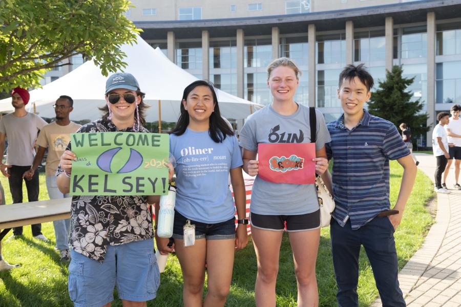 Four people casually stand in a row outdoors, two holding "Welcome to Olin" signs.