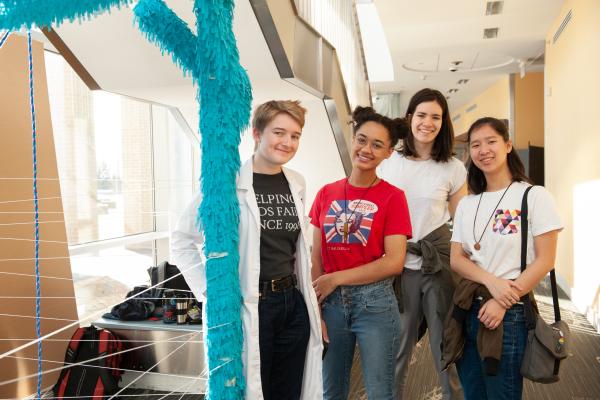 A photo of four students smiling while standing next to a tall blue tube 