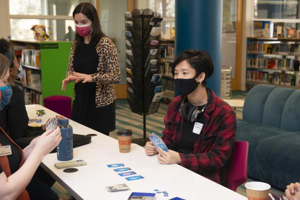 A student wearing a black face-mask plays cards with a friend in the Library.