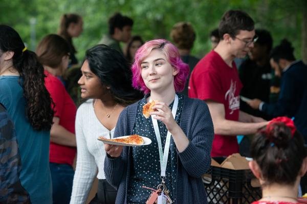 A student with pink hair eats a piece of pizza from a paper plate outside during an event on campus.