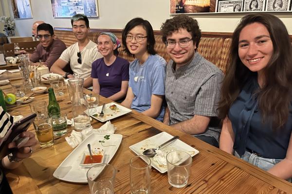 Six people look to the camera while seated next to each other on one side of a wood table.