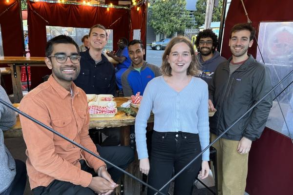 Six people look to the camera as they sit and stand around a bar table.