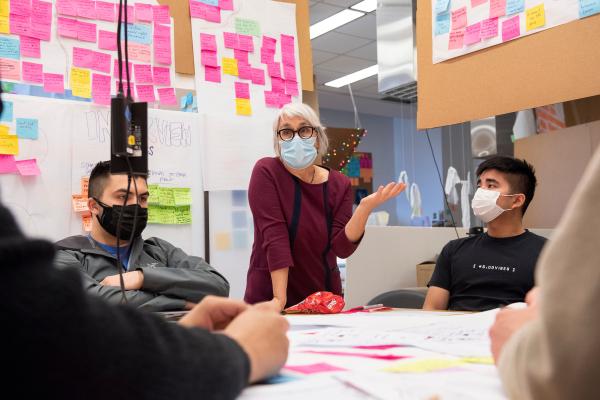 A teacher stands in front of a sticky note-filled wall to talk with students in a class.