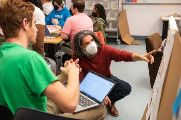 A teacher crouches down in a classroom to point to something as he talks with students.