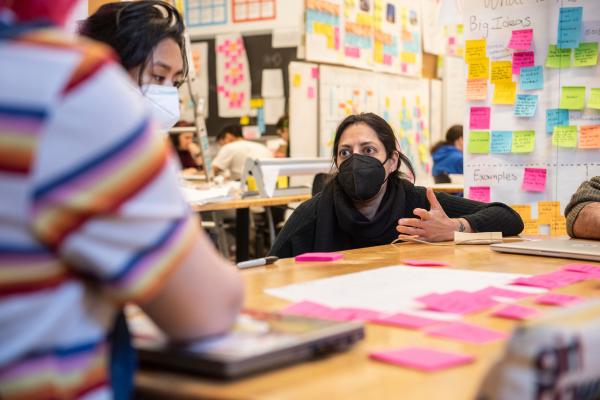 A teacher crouches down in a colorful classroom to talk with students.