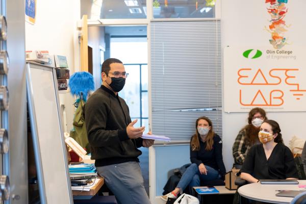 A teacher stands in front of a classroom holding a blue, square device.