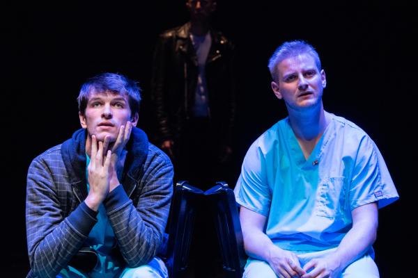 Two men sit on stage, bathed in blue light, with a person in shadow behind them.