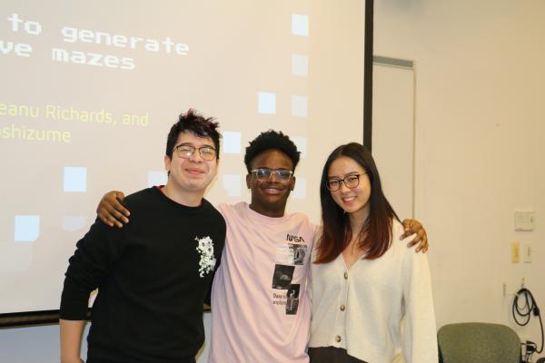 Three students pose in front of a presentation screen with arms around each other.