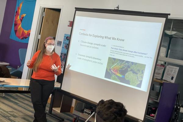 A women wearing a face mask and orange sweater presents in front of a large screen.