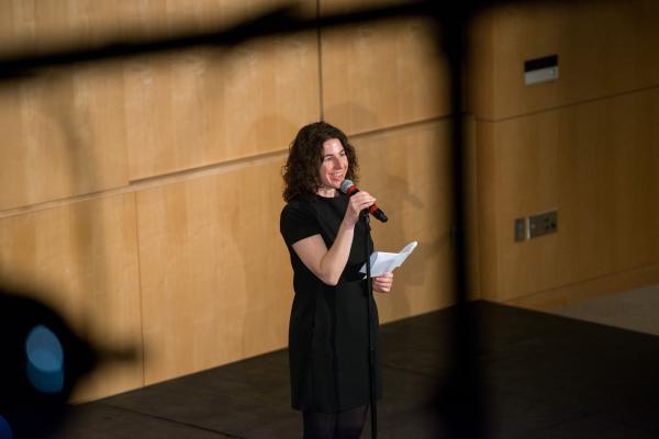 A woman in a black dress stands in front of an audience with a microphone