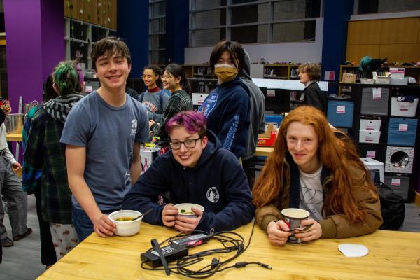Four students - two sitting, two standing - enjoy bowls of ramen in a library.