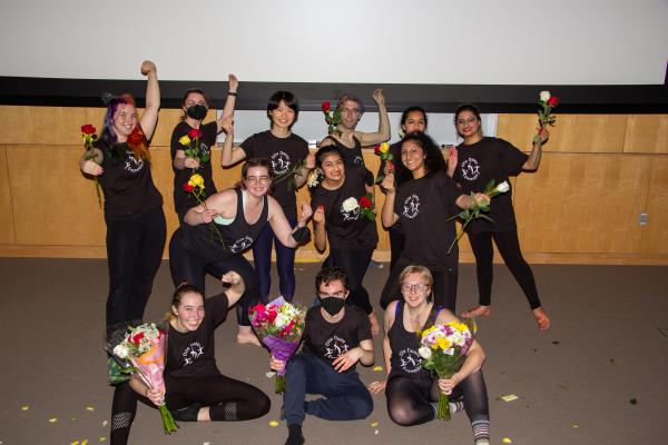 A medium-sized group of dance students pose for a photo wearing black shirts and holding flowers.