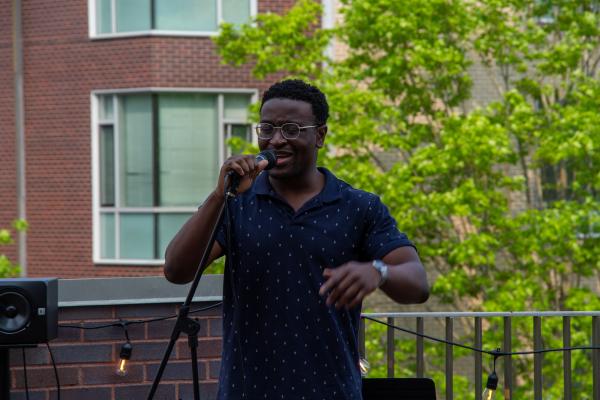 A young man wearing a dark shirt and glasses performs with mic in hand at the Art in the Oval event.