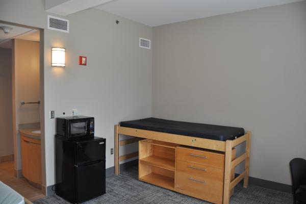 View of bed with under bed dresser and bookshelf next to microwave-fridge unit