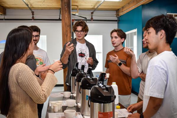 Students stand around a table with airpots with coffee. 
