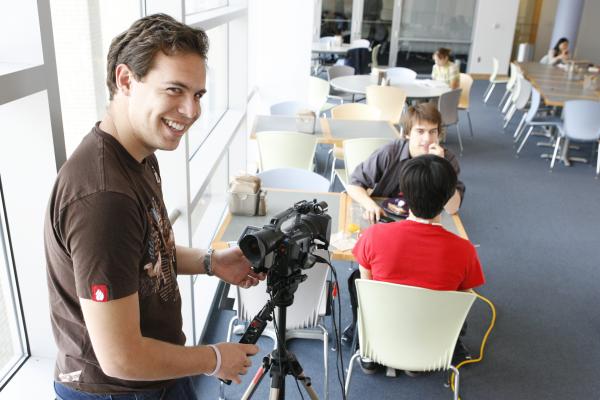 Kevin Tostado ’06 smiles with a camera as he films two students sitting down at a table. 