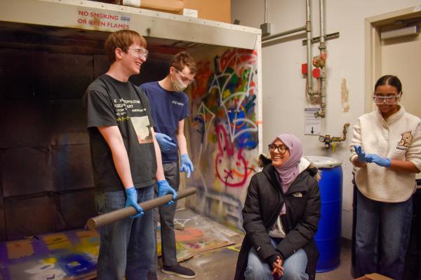 Four Olin Rocketry project team members wearing safety goggles stand together and smile at each other. One person holds a length of metal.